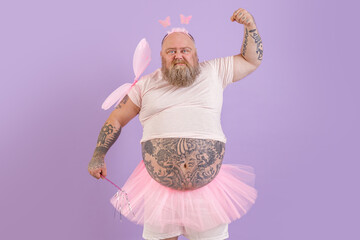 Funny brutal bearded obese man with tattoos wearing fairy costume with magic stick and wings shows...