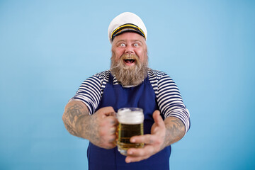 Happy mature fat man in sailor suit with apron and captain hat offers glass mug of fresh beer on...