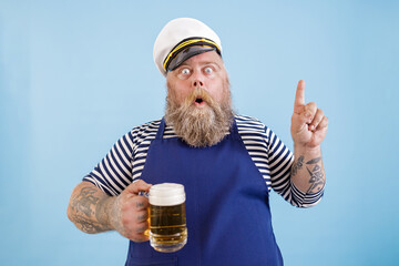 Emotional mature sailor man with overweight in apron and captainhat holds beer and gestures on light blue background in studio - obrazy, fototapety, plakaty