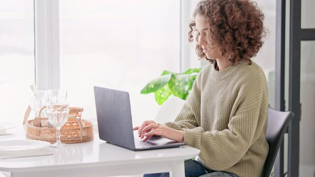 woman with curly hair in casual sweater sitting at the dinner table typing on laptop