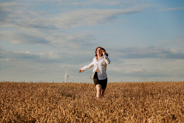 Naklejka na ściany i meble Happy woman in a white shirt runs in a wheat field on a summer day. Happiness and joy concept