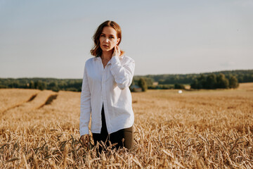 Naklejka na ściany i meble Portrait of a beautiful red-haired girl in a white shirt. She stands in a rye field on a sunny day. Calm facial expression. The concept of appeasement