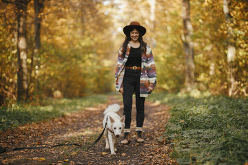 Fototapeta premium Cute dog walking on background of stylish woman traveler owner in sunny autumn woods. Traveling with pet, loyal companion. Young female hipster hiking with swiss shepherd white dog.