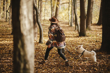Stylish woman in hat and with backpack walking with cute dog in sunny autumn woods. Young female traveler hiking with swiss shepherd white dog in beautiful forest. Travel with pet