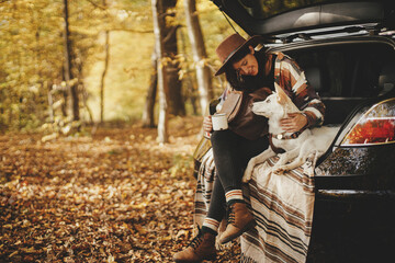 Stylish happy woman traveler sitting with cute dog in car trunk in sunny autumn woods. Travel and...