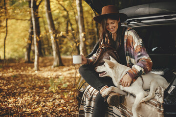 Stylish hipster woman with cup and backpack sitting with cute dog in car trunk in sunny autumn woods. Travel and road trip with pet. Space for text. Young female traveler hugging sweet white dog - obrazy, fototapety, plakaty