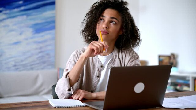 Focused confident smart african american curly haired girl in casual clothes, freelancer or student, working or studying remotely while sitting in living room at laptop and taking notes