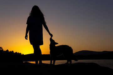 Silhouette of woman playing with a dog at sunset in front of a lake. Love for animals. Selective focus. Copy space.