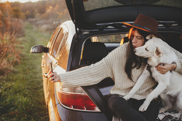 Happy stylish woman taking selfie photo with cute dog in car trunk in sunset light in field. Young hipster female using phone with sweet white dog. Autumn road trip with pet and travel