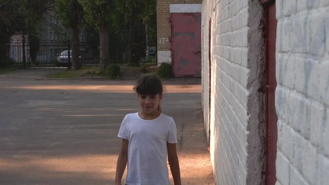 A young girl walks near the white wall. Urban area. White T-shirt. Evening. Walk down the street. School yard. Playground.