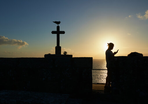 a man photographs the sunset with his smartphone next to a concrete cross where a seagull has perched