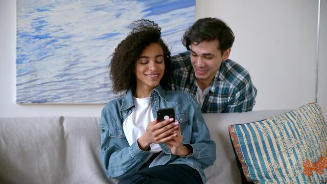 Millennial multiracial couple spends leisure time at home on the sofa, using a smartphone, browsing the Internet and social networks, laughing, the girl shows the guy something funny on the phone