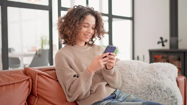 brunette sitting on the sofa holding smartphone messaging with friend weekend at home with fireplace cozy interior camera move smiling young woman with curly hair