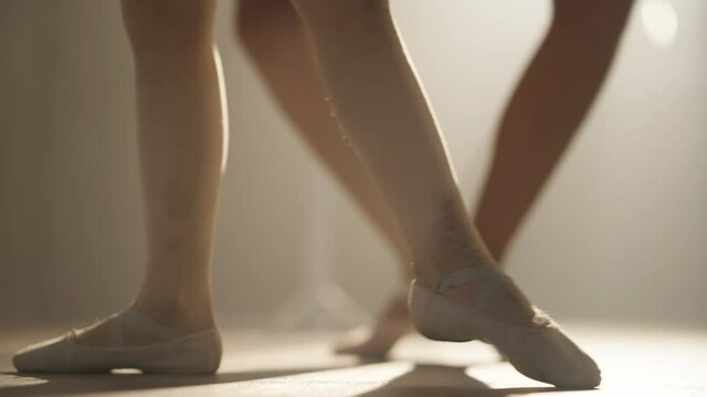 Legs of little ballerina in pointes in backlit fog as unrecognizable teacher adjusting tendu position. Slim unknown woman teaching ballet to talented motivated kid indoors in slow motion