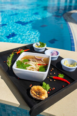 Noodle soup in luxury resort with swimming pool blurred in the background, garnished with herbs, red chili, garnish.