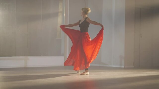 Motivated hard-working slim beautiful woman rehearsing ballet dance in slow motion in backlit fog. Caucasian ballerina in scarlet red skirt training performance in dancing studio in front of mirror