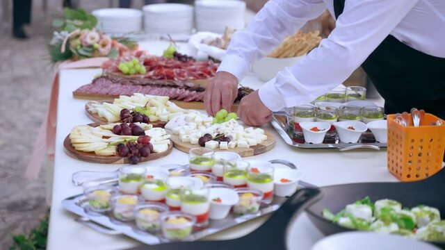 Restaurant waiters preparing buffet table for wedding party, servant putting cutlery for invited guests on the table. Silver trays and wooden boards full of meat snacks, appetizers and canapes