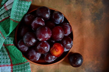 Black Cherry Plums and one slice in bowl and one pieces nearby next checkered towel on brown dark background. Top view