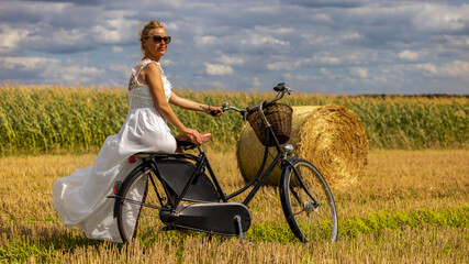 eine Frau mit einem weißen eleganten Kleid und einem Fahrrad auf einem Feld mit Strohballen