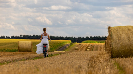 Naklejka na ściany i meble eine Frau mit einem weißen eleganten Kleid und einem Fahrrad auf einem Feld mit Strohballen