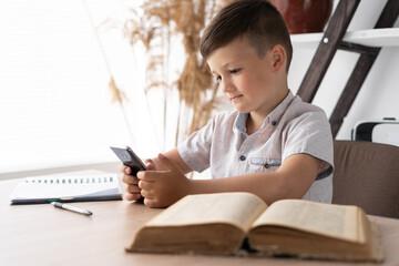 concentrated boy playing a game on the mobile phone while sitting at the table does homework. Gadget addicted student. Concept for online games or social network.