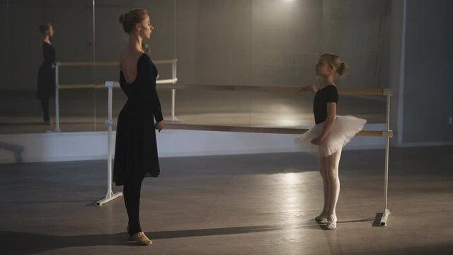Side view wide shot of slim woman and inspired girl rehearsing eleve at barre in ballet studio. Confident Caucasian teacher and diligent motivated student repeating ballroom movement