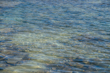 Meditative ripple of mountain lake. Beautiful relaxing background of blue green transparent water of glacial lake in sunlight. Sunny nature backdrop with many stones in clear water of glacier lake.