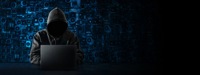 Hacker with a hidden face and with a laptop sits at the table, blue background. There is an...