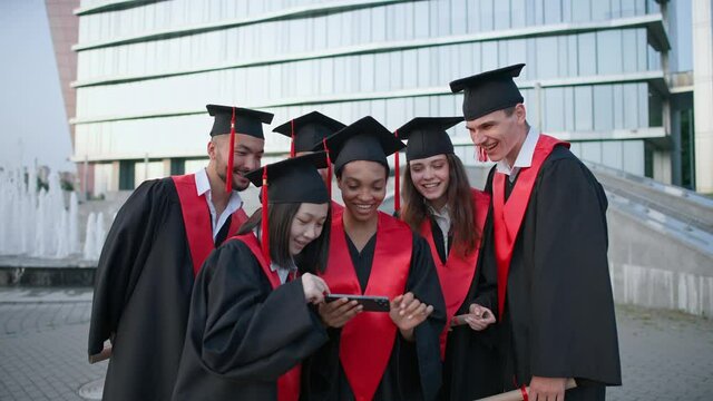Graduation at an international university, young people in graduate robes standing near the university and viewing photos on smartphone, positive emotions.