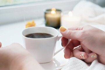 female hands holding white cup of coffee. autumn cozy morning, yellow manicure on nails. fall season candle on a windowsill.