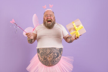Joyful middle aged man with overweight in fairy costume with wings holds gift box and magic stick...