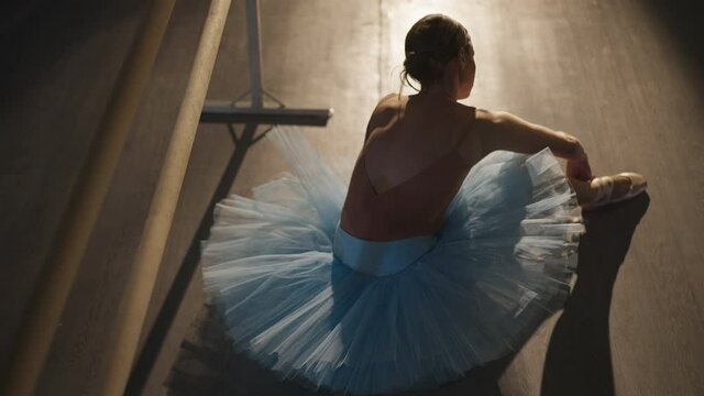 Back high angle view of slim talented hard-working ballerina in blue tutu tying laces on beige pointes indoors. Gorgeous graceful slender Caucasian woman getting ready for rehearsal in dancing studio