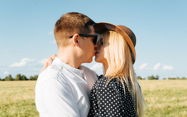 Kiss of beautiful young couple outdoors. During romantic walk in nature, man with glasses gently kisses his smiling girlfriend with closed eyes in straw hat. Love story