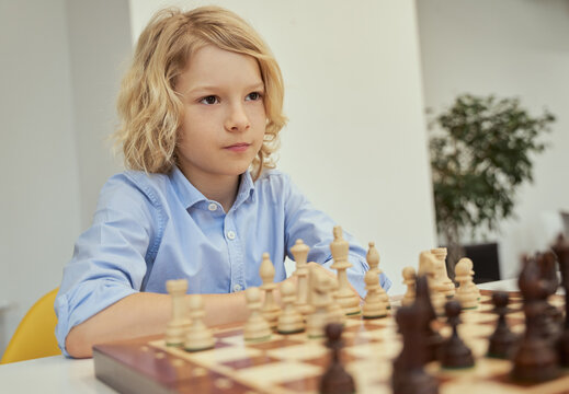 Portrait of calm caucasian boy in blue shirt sitting in the classroom and looking away while playing chess on the chessboard