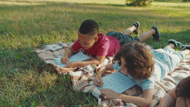Cheerful multiethnic diverse pupils lying on plaid, writing examination test outdoors. Handsome mixed race boy peeping into exam sheet of classmate while young teacher catching schoolboy on cheating