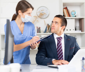 Fototapeta premium Young woman professional medical worker monitoring state of health of businessman working in office. Pandemic precautionary concept