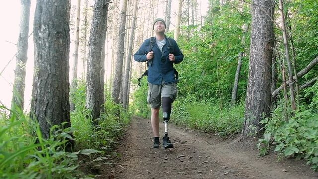 Disabled man with prosthetic limb with backpack walks through forest overcomes path and enjoys. Traveling on foot along forest road and active lifestyle for people with disabilities. active hiking