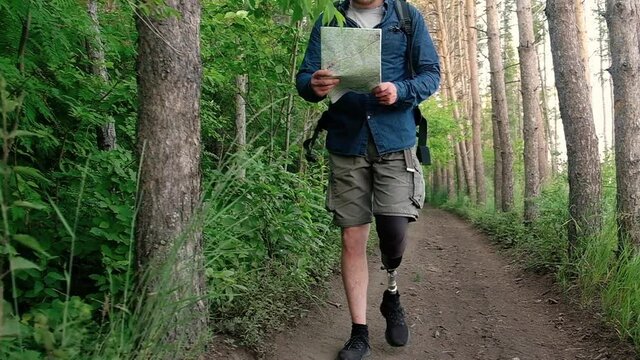 Disabled tourist with prosthetic leg walks along pedestrian path in forest and uses map for navigation. man with backpack is traveling along forest paths. Active weekend outside. Hiking in nature