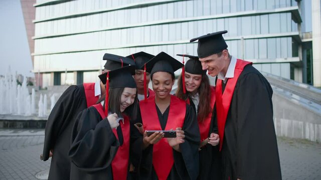 Graduation at an international university, young people in graduate robes standing near the university campus and viewing photos on smartphone, positive emotions.