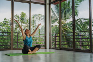 An attractive 40-year-old middle-aged woman practices yoga in a panoramic window room overlooking...