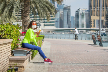 Fototapeta premium Asian woman sitting on a bench in the Dubai Marina area and wears a medical protective mask due to the coronavirus pandemic and uses a smartphone