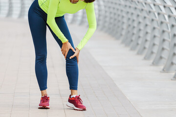 Young woman was running along the embankment and suddenly felt a sharp pain in the knee joint due to a dislocation or rupture of the meniscus - obrazy, fototapety, plakaty