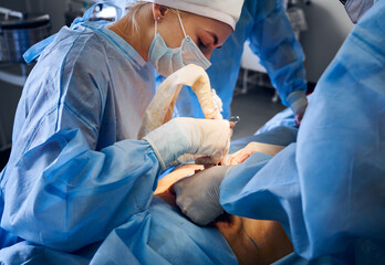 Doctors in blue uniforms performing surgical operation in operating room. Plastic surgeon and...
