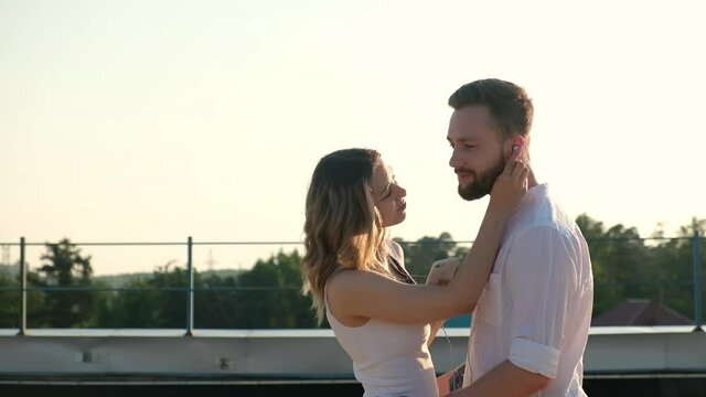 Young Caucasian lovers, woman and man standing on roof and hugging. beautiful girl puts on headphones to boyfriend and listens to music together. couple of people on roof at sunset in summer enjoying