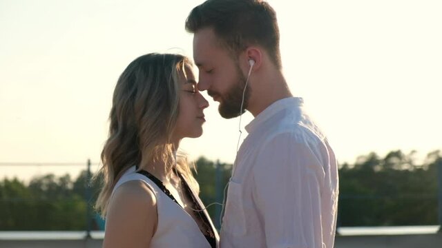Young Caucasian lovers woman and man are standing on roof and hugging and listening to music together with headphones. couple of people on roof in evening at sunset in summer enjoy vacation together.