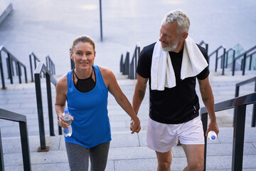 Fototapeta premium Attractive middle aged couple of sportive man and woman looking happy, holding hands while walking up the stairs after training together in the city