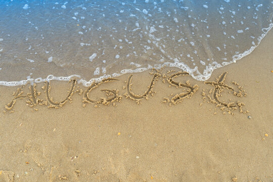 The word August is written on the sand of a beach by the sea. Washed off by a wave. The inscription disappears. End of summer concept. Back to school