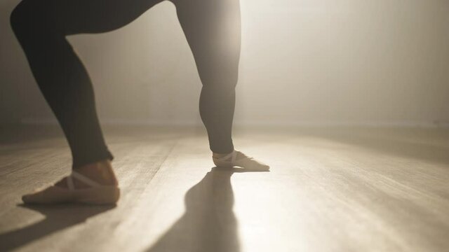 Camera moves down as slim graceful confident ballerina rehearsing in backlit fog indoors. Professional Caucasian female ballet dancer performing on stage in spotlight smoke