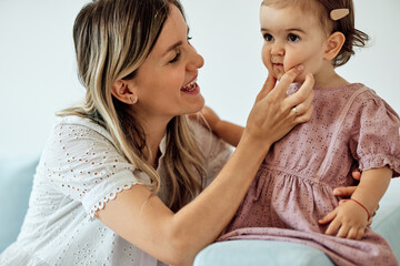 Young beautiful mother sitting at home with her baby girl and stiffing its chubby cheeks.