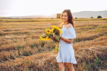 Young happy brunette on sunset walking thru the meadow with handful of sunflowers